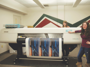Roberta Figolia and Paige Lavery, both sophomores in the Graphic Media Management Program. demonstrate how to use the new large format printer, that was in the box.